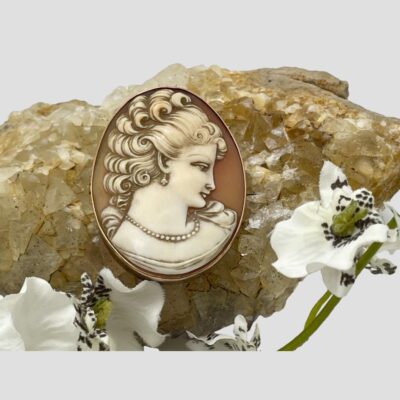 Main photo of brooch displayed resting on top of a rock of natural quartz crystal of a light sand colour and a small spray of white orchids laid across the bottom. The lady is looking right and down with a very contented look on her face. Her curly hair is trussed up in a loose bun with curls cascading down the back of her neck and she is also wearing pearls which sit tightly around her collar bone and a earring is visible in her ear.