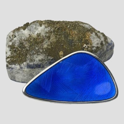 Main photo of brooch leaning up against a natural rock which houses the lapis but not showing the lapis side (although some blue coming thru the rock) The brooch is shown with the longest edge on the surface and the shortest edge to the right. This blue enamel is slightly darker like royal blue. There are multiple triangles sections within the triangle form each defined with horizontal lines which make them stand out.