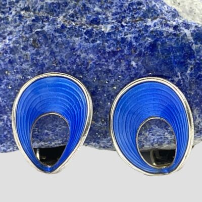 Main photo of earrings displayed in front of a piece of natural smooth rock of blue lapis lazuli rock with the earring fronts forward facing. The blue enamel on the earrings are of a more vibrant colour and stand out well against the blue of the lapis. The shape of the earring front is like a parachute with a concave dip in the middle and framed within a sterling silver border.