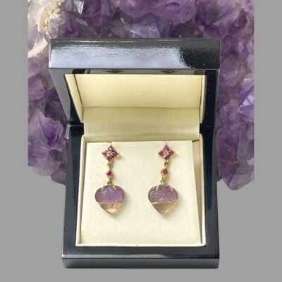 Main photo of earrings displayed inside a box which is leaning up against a rock of natural amethyst. Ametrine is amethyst and citrine stones together and a natural occurrence. These stones have the purple amethyst area to the top half of the heart shaped stone and carved with the pale yellow citrine part at the lower point. The 18 karat gold the stone is set on has prongs situated exactly at point the 2 stone colours separate enhancing the stones look & quality. The heart is attached to a short 18 karat gold chain which has a square cut ruby to the centre. The top of the earrings in stud form also have 4 rubies laid out in a floral pattern. Each piece of these earrings compliment each other perfectly.