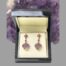 Main photo of earrings displayed inside a box which is leaning up against a rock of natural amethyst. Ametrine is amethyst and citrine stones together and a natural occurrence. These stones have the purple amethyst area to the top half of the heart shaped stone and carved with the pale yellow citrine part at the lower point. The 18 karat gold the stone is set on has prongs situated exactly at point the 2 stone colours separate enhancing the stones look & quality. The heart is attached to a short 18 karat gold chain which has a square cut ruby to the centre. The top of the earrings in stud form also have 4 rubies laid out in a floral pattern. Each piece of these earrings compliment each other perfectly.