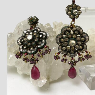 Main photo of earrings with one hanging from a display stand to the right of photo and other laid on top of a rock of natural quartz crystal. The earrings are sparkling beautifully in the light. There is a small daisy shaped area housing the post to the top with a larger daisy shaped section hanging from the smaller one. Along the bottom of the larger daisy are 5 small gilt rings with small amethyst stones & golden balls hanging on each and from the central one of these a water drop shaped deep pink stone bead. The earrings are truly the essence of Bollywood past!