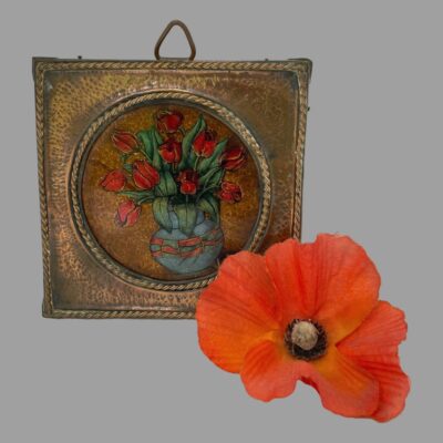 Main photo of picture displayed upright and forward facing. The tulips are red and inside a light blue bulbous vase with red accents. The hook for hanging the picture is poking out from the top centre and there is a single red poppy laid to the right of the picture.