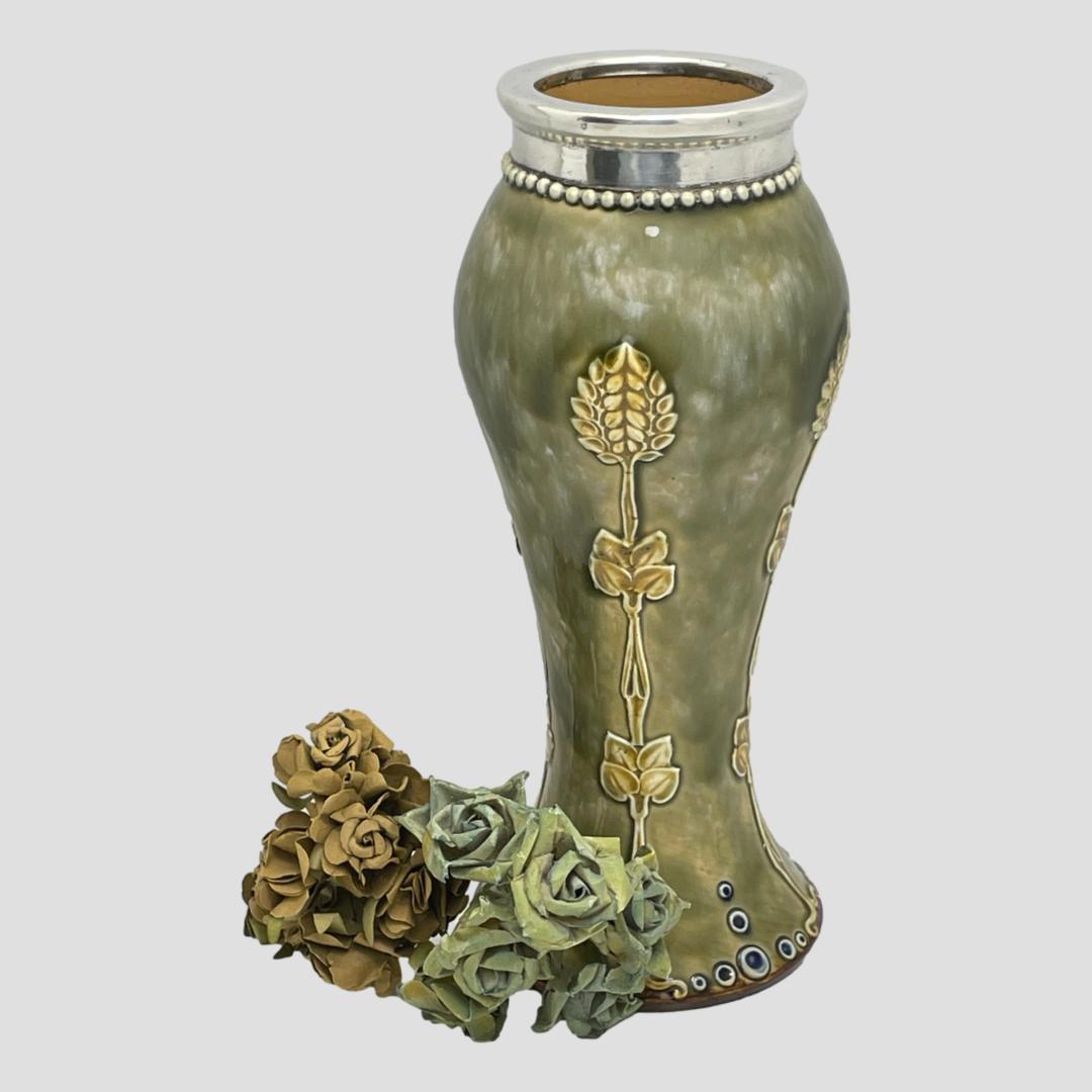 Main photo of vase seen from an almost eye level with just a slight peek inside at the natural unglazed stoneware. One of the applied Art Nouveau decorations which looks like a wheatsheaf is on show to the side with the small blue bubble like decoration along the base rim. The silver rim at top finishes the vases elegance. There is a small posy of light rust and pale blue/green roses to the left of the vase.