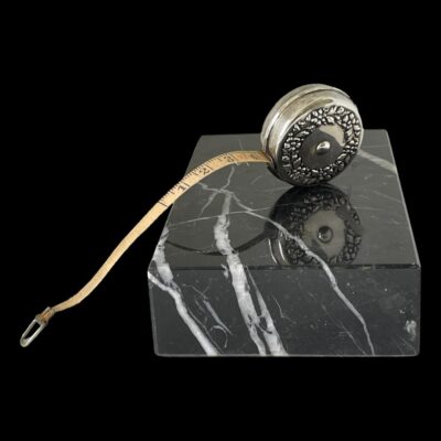 Main photo of tape measure shown with some tape sticking out . The tape measure is balanced upright on top of a slab of black marble with white line inclusions and the 4 inches of sticking out tape is to the left. The side of tape measure on display is the non retractable button side which is plain silver.
