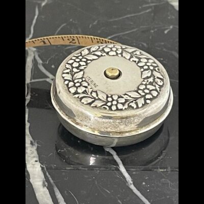 Photo of tape measure laid flat on top of the black marble with the gilt silver retractable button visible to the centre. There is a small bit of tape sticking out. The Sterling mark is visible to the inside of garland and on the left.