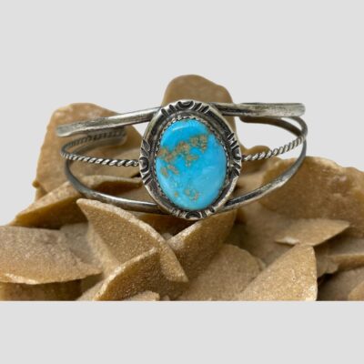 Main photo of cuff bracelet displaed on top of a rock of natural desert sand rose with the cuff front and turquoise forward facing. The turquoise is set on a frame of sterling silver with the frame of the cuff in 3 lines 2 smooth silver to the top and bottom and a twist line through the centre. The silver frame around the turquoise has short lines to the north, east, south and west of the turquoise representing the 4 winds perhaps.