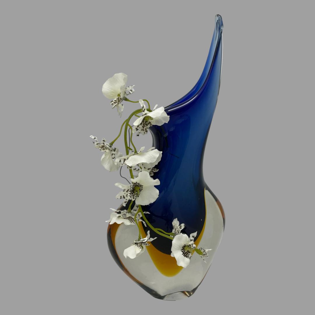 Main photo of vase seen slightly angled to the side so one wide side is turned a little to the right. The vase has an uneven top in blue with one side raised up to a point and other rounded and overhanging almost like a fly catcher pitcher plant. The lower part of the vase is bulbous in clear glass. The blue upper glass part merges into the clear glass at the base with amber coloured glass within the clear glass and at the bottom of the blue. There is a small spray of white orchids hanging down from the lower lipped left side of the vase,