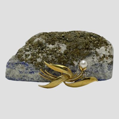 Main photo of brooch leaning against a small rock with gold and blue inclusions. The brooch is shown forward facing and has a 3 leaves in the lower area spread in all directions with 3 stems visible to the left. A single stem rises up to the centre of top right which holds the single pearl and the stems with small diamonds on the ends are to the left and right of the pearl stem, slightly lower.