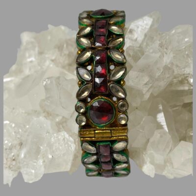 Main photo of bracelet shown vertically upright with a rock of natural quartz crystal through the middle holding it up. The centre row of the bracelet houses the dark pink stones with clear glass circles and petals on either side. Each area with a glass stone piece is enhanced with an outer painted frame of bright green enamel which along with the gilt, makes this bracelet truly stand out.