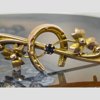 Close up photo of the horseshoe area of the brooch. There is a bar with scrolled ends which sits diagonally (not straight) The horseshoe is mounted with one end this side of the bar and other side behind the bar. The single sapphire is set on the bar in an area central to the horseshoe and finally finished with a single stemmed floral spray coming from the sapphire to the left and right.