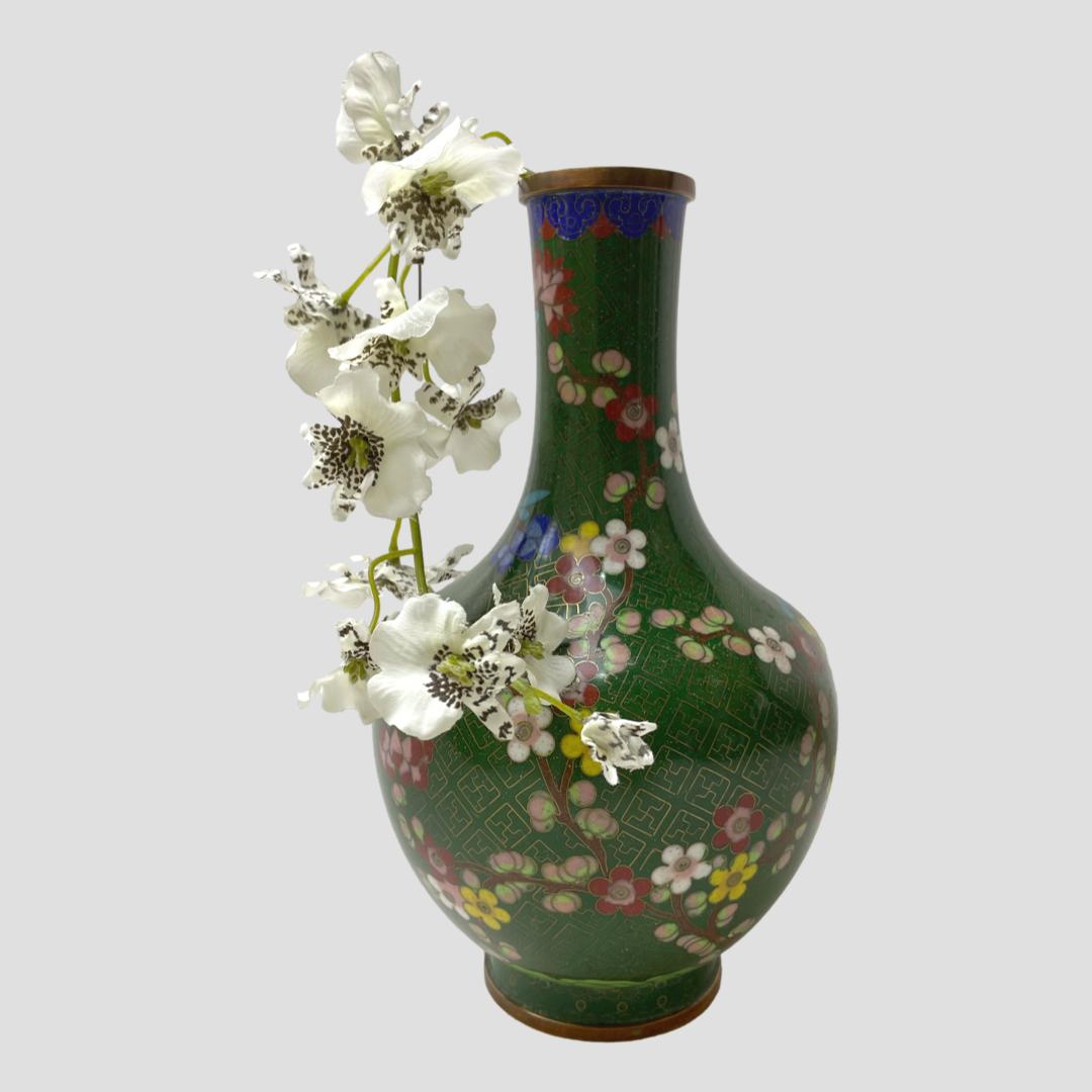 Main photo of vase seen from an eye level with the side with most blossoms on display. There is a single spray of small white orchids. hanging down on the left hand side. The blossoms in red, pink, yellow and blue with green foliage stands out beautifully against the racing green coloured background.