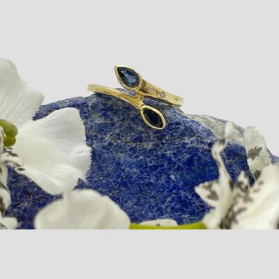Main photo of ring displayed on top of a rock of natural lapis lazuli with small white orchids framing the base of the lapis. The ring front is forward facing. The pear cut sapphire is to the top with the small round cut diamonds to the side band.