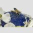 Main photo of ring displayed on top of a rock of natural lapis lazuli with small white orchids framing the base of the lapis. The ring front is forward facing. The pear cut sapphire is to the top with the small round cut diamonds to the side band.