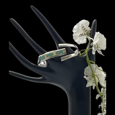 Main photo of bracelet shown displayed hanging on the fingers of a black display hand and seen with bracelet front forward facing. The single band end housing the 3 small panels of turquoise is to the left and the split band end to the right. The turquoise panels are rectangular and run consecutively next to each other. There is also a spray of small white orchids hanging down off the stands thumb.