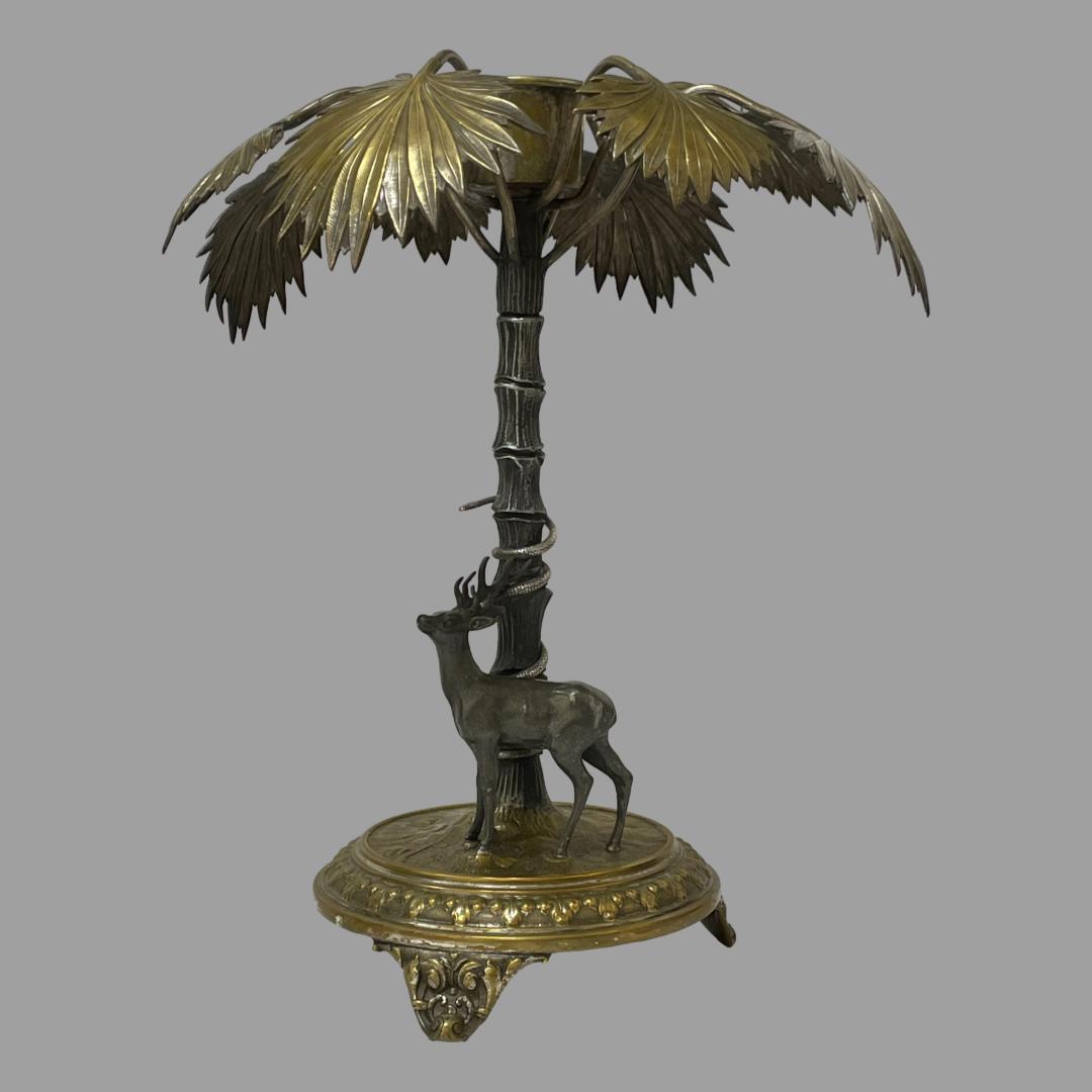 Main photo of the candlestick from an eye level with the stag facing to the left. The snake can be seen coiled to the palm trees trunk behind the stag and also facing the same direction. The fan palm fronds appear like an umbrella sheltering the 2 creatures.