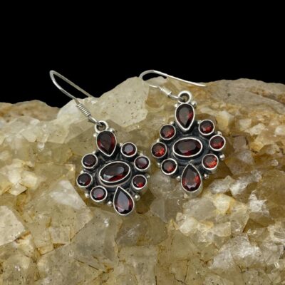 Main photo of earrings displayed side by side on top of a rock of natural river quartz. The quartz is of a slightly pale orange colour which brings out the beautiful red of the garnets perfectly. The garnets are set out with an oval cut stone horizontal at the centre with a pear cut garnet with pointed ends facing inwards to the top and bottom of central garnet. To the immediate left and right of the central oval garnet there is a line of 3 vertically set small round cut garnets.