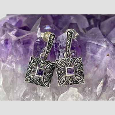 Main photo of earrings displayed side by side and hanging from a rock of natural amethyst. The top area with posts attached are like a half hoop but wider at top finishing almost in a point at bottom which curves backwards like a hook. Attached to this upper piece is a square of silver with marcasite and a square cut amethyst to the centre. Petal shapes extend from the 4 corners of the central square like a flower.