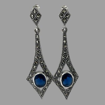 Main photo of earrings displayed hanging. There is a small diamond shape at the top where the posts are and an elongated diamond shape (long at top) with the blue cubic zirconia housed between the 3 lower diamond points. The cubic zirconia is a very beautiful shade of blue like a lagoon blue, not dark not light just right.