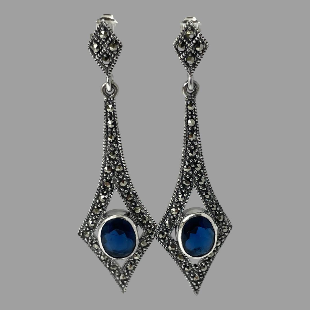 Main photo of earrings displayed hanging. There is a small diamond shape at the top where the posts are and an elongated diamond shape (long at top) with the blue cubic zirconia housed between the 3 lower diamond points. The cubic zirconia is a very beautiful shade of blue like a lagoon blue, not dark not light just right.