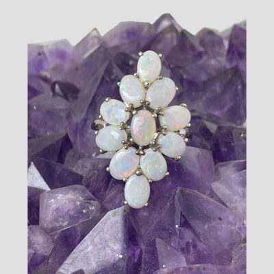 Main photo of ring displayed on top of a rock of amethyst with ring front facing forward. There is an opal cabochon to the centre vertically set with 8 opals surrounding it, shorter sides in so like flower petals and an opal to the top and bottom. The colours are subtle but very visible.