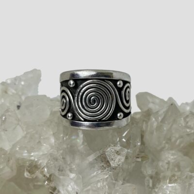 Main photo of ring displayed on top of a rock of natural quartz crystal with the ring front forward facing. The large swirl is to the centre with 2 small swirls coming off it to either side. There are 4 dots spread out top left and right and bottom left and right of large swirl. There is a band of plain silver to the top and bottom edge of the ring.
