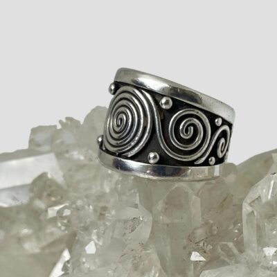 Photo of ring still displayed on top of a rock of quartz crystal with the ring front facing towards the left. The main large swirl is partially visible with the smaller swirl coming off it and a smaller swirl coming off the second swirl. There are dots evenly spread around the ring.