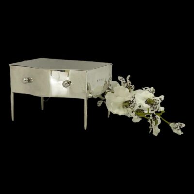 Main photo of jewellery box shown at eye level and closed with the front facing slightly to the left. Two small silver knobs are visible to the left and right of the frontage with the thin silver legs also visible. There is a sprig of small white orchids to the right.