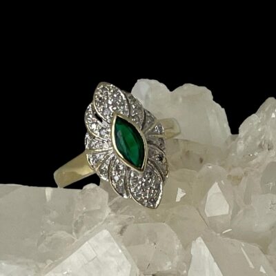 Main photo of ring displayed on top of a rock of natural quartz crystal with the ring front forward facing. The ring is on a slight slant raised a little on the right side. The emerald is a beautiful green shade in oval cut with a thin frame of 14 karat gold around it. The diamonds are set on a scallop edged frame around the emerald. 2 petal shaped areas are above and below the emerald housing 6 diamonds each. On either side of these petal shapes are strips either side with 3 diamonds in a line in each strip, 2 diamonds in a strip each to either side of these and finally a single diamond immediately to the left and right of the emerald. Each area of diamonds is separated by a thin line of gold.
