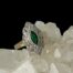 Main photo of ring displayed on top of a rock of natural quartz crystal with the ring front forward facing. The ring is on a slight slant raised a little on the right side. The emerald is a beautiful green shade in oval cut with a thin frame of 14 karat gold around it. The diamonds are set on a scallop edged frame around the emerald. 2 petal shaped areas are above and below the emerald housing 6 diamonds each. On either side of these petal shapes are strips either side with 3 diamonds in a line in each strip, 2 diamonds in a strip each to either side of these and finally a single diamond immediately to the left and right of the emerald. Each area of diamonds is separated by a thin line of gold.