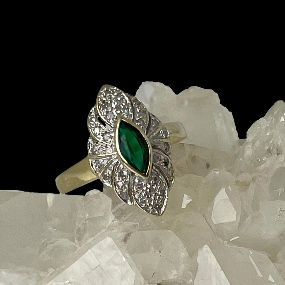 Main photo of ring displayed on top of a rock of natural quartz crystal with the ring front forward facing. The ring is on a slight slant raised a little on the right side. The emerald is a beautiful green shade in oval cut with a thin frame of 14 karat gold around it. The diamonds are set on a scallop edged frame around the emerald. 2 petal shaped areas are above and below the emerald housing 6 diamonds each. On either side of these petal shapes are strips either side with 3 diamonds in a line in each strip, 2 diamonds in a strip each to either side of these and finally a single diamond immediately to the left and right of the emerald. Each area of diamonds is separated by a thin line of gold.