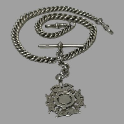 Photo of the chain displayed on a flat surface. The fob is in the centre foreground, the bail is above it off a short chain. To the left and right of the bail are the 2 graduating chains, the right hand one is swirled inward with dog clip at the centre and other is curved above and over this with the dog clip clasp ending at the right. The T-Bar is above the bail and laid horizontally.